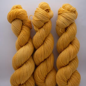 May include: Three skeins of golden yellow yarn are displayed against a white background. The yarn is tightly twisted into a rope-like shape, with a soft, textured appearance. The color is a vibrant, sunny yellow, perfect for knitting or crocheting projects.