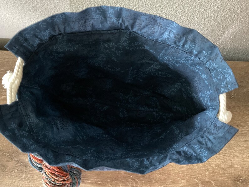May include: A handmade tote bag with a navy blue floral patterned interior. The bag has a white rope handle and a decorative tassel at the bottom. The bag's exterior is not visible in this image.