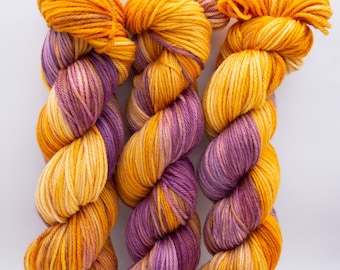 May include: Three skeins of hand-dyed yarn in a vibrant color palette. The yarn features a blend of golden yellow, orange, and purple hues, creating a visually appealing gradient effect. Ideal for knitting or crochet projects.