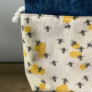 May include: A fabric storage bag with a bee and honeycomb pattern. The bag features a white background with yellow honeycomb shapes and black bees. The top edge is navy blue with a floral design, and a white rope drawstring is visible.