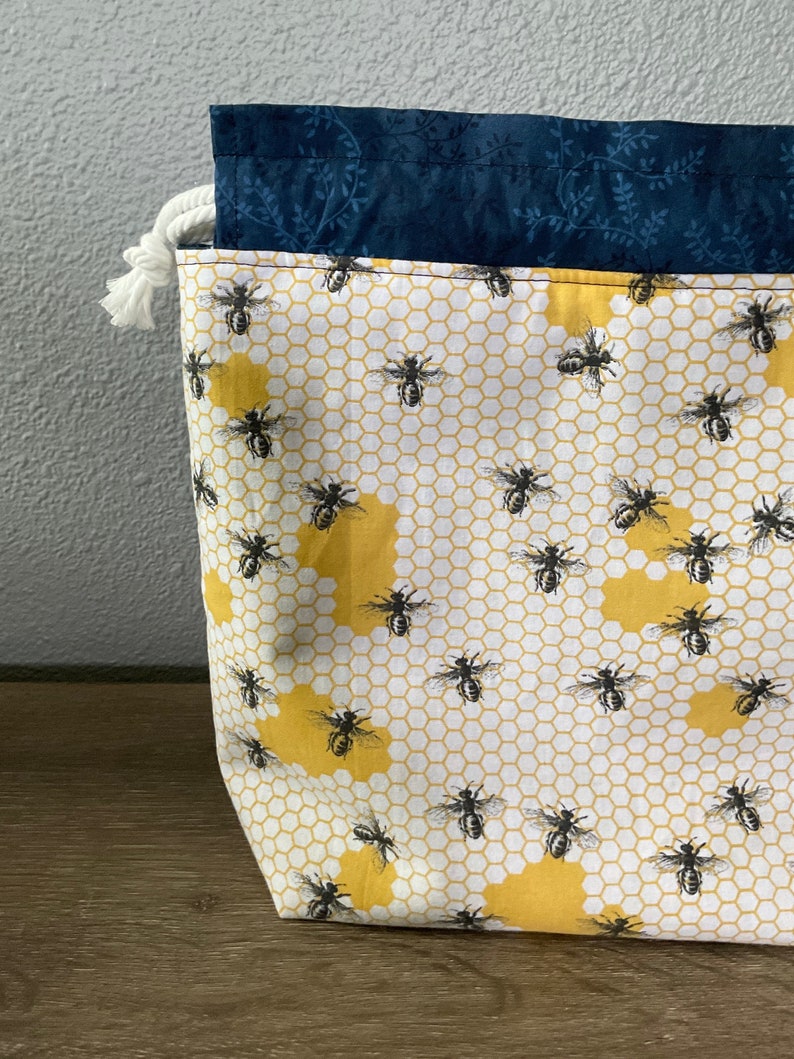 May include: A fabric storage bag with a bee and honeycomb pattern. The bag features a white background with yellow honeycomb shapes and black bees. The top edge is navy blue with a floral design, and a white rope drawstring is visible.