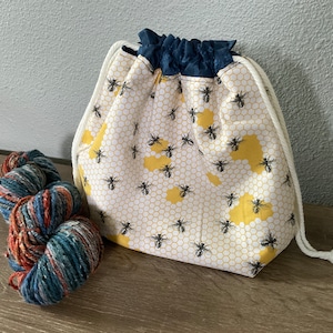 May include: A drawstring project bag with a bee and honeycomb pattern, next to a skein of yarn. The bag features a white background with a yellow honeycomb design and black bees. The top is navy blue.