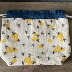 May include: A drawstring bag with a white background, featuring a pattern of yellow honeycomb shapes and black bees. The top edge is a dark blue fabric, and the drawstring is white.
