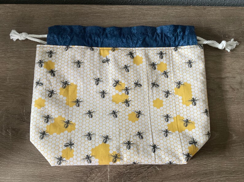 May include: A drawstring bag with a white background, featuring a pattern of yellow honeycomb shapes and black bees. The top edge is a dark blue fabric, and the drawstring is white.