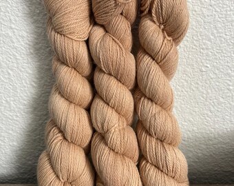 May include: Three skeins of light brown yarn are arranged vertically against a white wall. The yarn is tightly twisted, creating a textured appearance. The color is a warm, neutral tone, suitable for various knitting or crochet projects.
