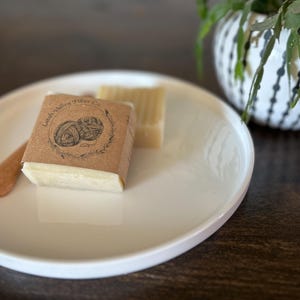 May include: A white ceramic plate holds two bars of soap, one wrapped in brown paper with the text "Latah Valley Fiber Co." and a graphic of yarn. A small wooden spatula rests on the plate. A decorative white and black pot with a plant is in the background.