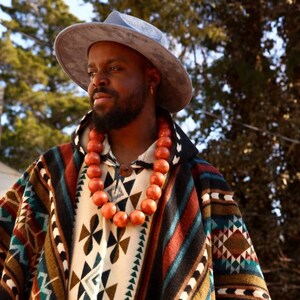 May include: A person wearing a blue hat and a colorful patterned poncho with a long orange beaded necklace.