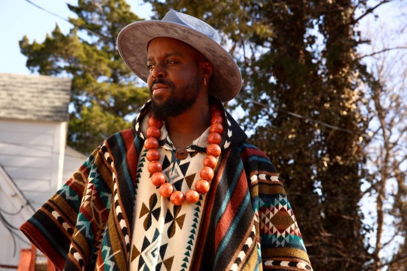 May include: A person wearing a blue hat and a colorful patterned poncho with a long orange beaded necklace.