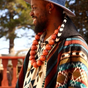 May include: A man wearing a blue hat and a colorful patterned poncho with a long necklace of wooden beads.