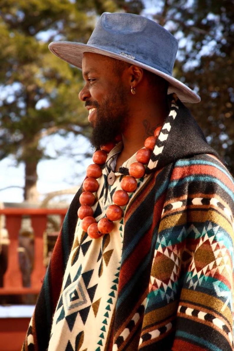 May include: A man wearing a blue hat and a colorful patterned poncho with a long necklace of wooden beads.