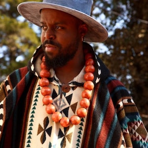 May include: A man wearing a blue hat and a colorful patterned poncho with a long orange beaded necklace.