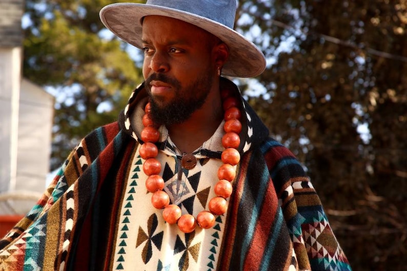 May include: A man wearing a blue hat and a colorful patterned poncho with a long orange beaded necklace.