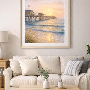 May include: A framed print of a pier extending into the ocean at sunset. The sky is a blend of orange and yellow hues, reflecting on the water. The pier has small buildings and an American flag. The artwork is displayed above a beige sofa.