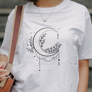 May include: White t-shirt with a black crescent moon design. The moon is adorned with delicate floral details and a small star. The design is surrounded by small stars and dots.