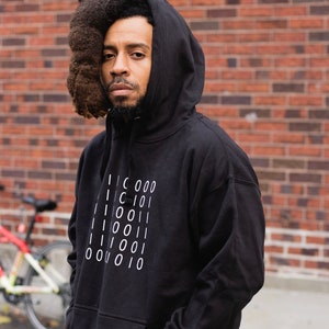 May include: Black hoodie with white binary code design. The hoodie has a front pocket and a hood. The person in the image is wearing the hoodie and dark jeans. A red and white bicycle is visible in the background.