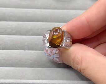 May include: A silver ring with an oval, amber-colored gemstone. The ring is held up, with other rings visible in the background. The gemstone has a fiery, translucent appearance, and the silver setting has a textured design.