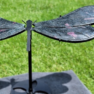Puede incluir: Una escultura de libélula de metal negro con alas detalladas y un cuerpo delgado. Las alas tienen una apariencia texturizada con toques de rosa y están muy extendidas. La escultura está montada sobre un soporte negro, sobre un fondo de hierba verde.