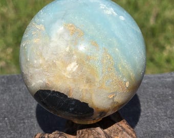 May include: A polished, spherical gemstone with a blend of blue, tan, and black hues. The sphere rests on a natural, dark brown wooden base. The text "Craven Crystals" is visible at the bottom.