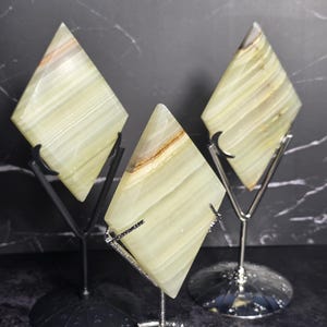 May include: Three diamond-shaped onyx stones in shades of green and cream, displayed on decorative metal stands. The stones have natural veining and are polished to a smooth finish. The stands are black and silver.