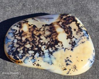 May include: A polished, oval-shaped gemstone with a creamy yellow base, adorned with intricate patterns of dark brown and blue. The stone's surface is smooth and glossy, with the text "Graven Crystals" at the bottom left.
