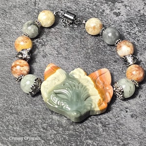 May include: A beaded bracelet featuring a carved fox head pendant. The bracelet includes round, multi-colored stone beads and silver-toned accents. The fox pendant is primarily green, orange, and cream-colored.