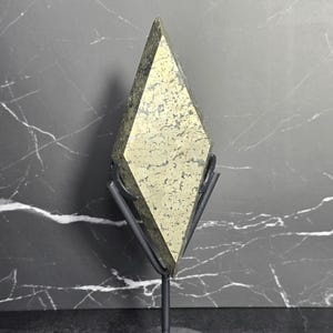 May include: A pyrite crystal in a diamond shape, mounted on a dark metal stand. The crystal has a metallic gold color with a textured surface. The stand is a simple, minimalist design, and the background is a dark gray with white veining.
