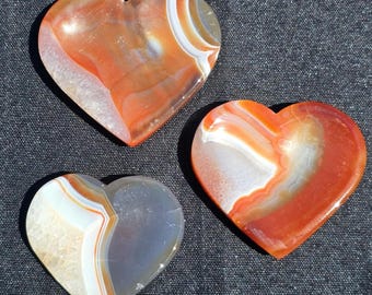 Puede incluir: Tres piedras de ágata en forma de corazón con bandas naranjas, blancas y grises. Las piedras tienen una superficie pulida y están dispuestas sobre un fondo gris oscuro. El texto "Craven Crystals" es visible en la esquina inferior izquierda.