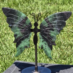 May include: A butterfly sculpture with green and black wings, mounted on a metal stand. The wings have a marbled effect, and the sculpture is set against green grass. The base of the stand is a silver disc. The text "Craven Crystals" is visible.