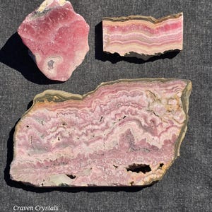 May include: Three pieces of raw rhodonite crystals in shades of pink, with layered patterns and natural textures. The crystals vary in size and shape, showcasing the stone's unique veining and color variations. The image is taken on a gray surface.