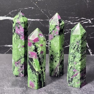 May include: Four polished zoisite crystal towers. The crystals are primarily green with vibrant pink and black inclusions. Each tower has a pointed, faceted top. The crystals are displayed on a gray surface with a black and white background.