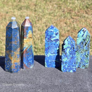 以下が含まれることがあります： 青、緑、茶色の色合いの、磨かれたアズライトクリスタルオベリスクのコレクション。 オベリスクはサイズと形が異なり、丸みを帯びたものと尖ったものがあります。 「Craven Crystals」の文字が見えます。 高さ約5〜10cmです。