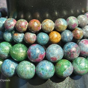 May include: A multi-strand bracelet composed of round, polished gemstone beads in a vibrant mix of blue, green, and magenta hues. The beads feature natural mineral inclusions and a glossy finish, strung together on a flexible cord. Text reads Craven Crystals.
