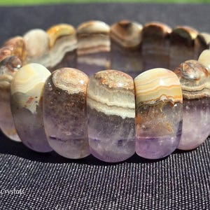 Puede incluir: Una pulsera elástica compuesta por cuentas de piedra rectangulares pulidas que presentan un degradado de amatista púrpura translúcida en la base, pasando a capas veteadas de color crema, beige y marrón. El texto Graven Crystals es visible.