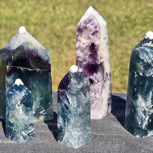 May include: Five fluorite crystal obelisks in shades of purple, blue, and green. The crystals have a polished, smooth surface and are topped with a small white cap. The obelisks vary in size and shape, standing on a dark surface.