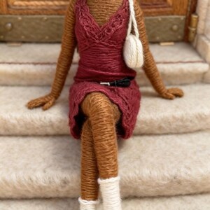 May include: A handcrafted doll made from brown yarn, dressed in a burgundy dress, with a white purse and black shoes. The doll has brown hair and is seated on a staircase. The doll's features are carefully detailed, demonstrating the artistry of the craft.