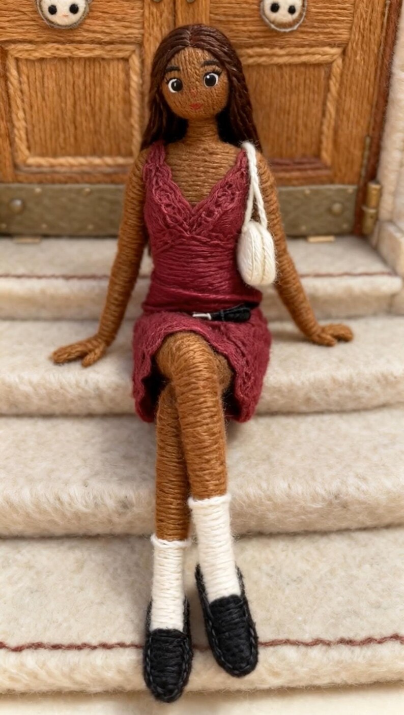 May include: A handcrafted doll made from brown yarn, dressed in a burgundy dress, with a white purse and black shoes. The doll has brown hair and is seated on a staircase. The doll's features are carefully detailed, demonstrating the artistry of the craft.