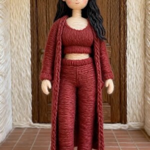 May include: A handmade doll dressed in a red knitted ensemble: a crop top, trousers, and a long cardigan. The doll has black hair and wears white slippers with cherry motifs. The doll is positioned in front of a brown wooden door.