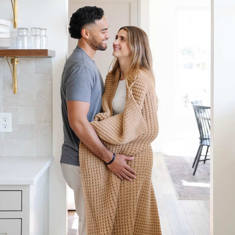 May include: A couple stands close, the woman draped in a tan waffle-knit blanket. The man wears a grey t-shirt and light trousers. The setting is a bright, modern interior with a kitchen visible in the background.