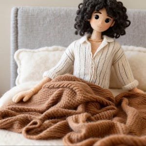 May include: A handcrafted doll with dark, curly hair, dressed in a white shirt with black stripes, seated on a bed with a brown knitted blanket. The doll has a calm expression and is surrounded by soft pillows.