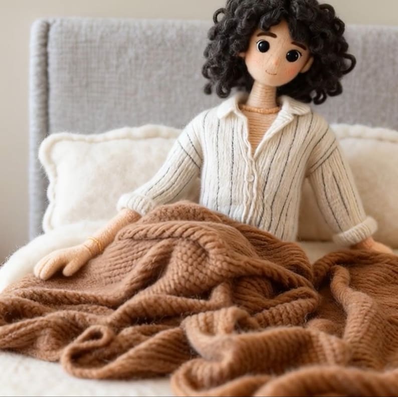 May include: A handcrafted doll with dark, curly hair, dressed in a white shirt with black stripes, seated on a bed with a brown knitted blanket. The doll has a calm expression and is surrounded by soft pillows.