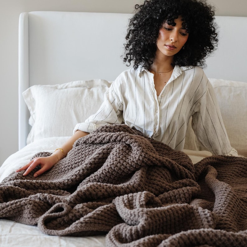 May include: A brown textured blanket with a waffle weave, draped over a bed. The bed has white linen bedding and a white headboard. A person wearing a striped shirt is sitting on the bed. The blanket is made of a soft material.