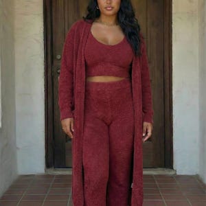 May include: A burgundy three-piece loungewear set, including a crop top, high-waisted trousers, and a long cardigan. The model is wearing white slippers with a cherry design. The outfit is made of a soft, textured material.