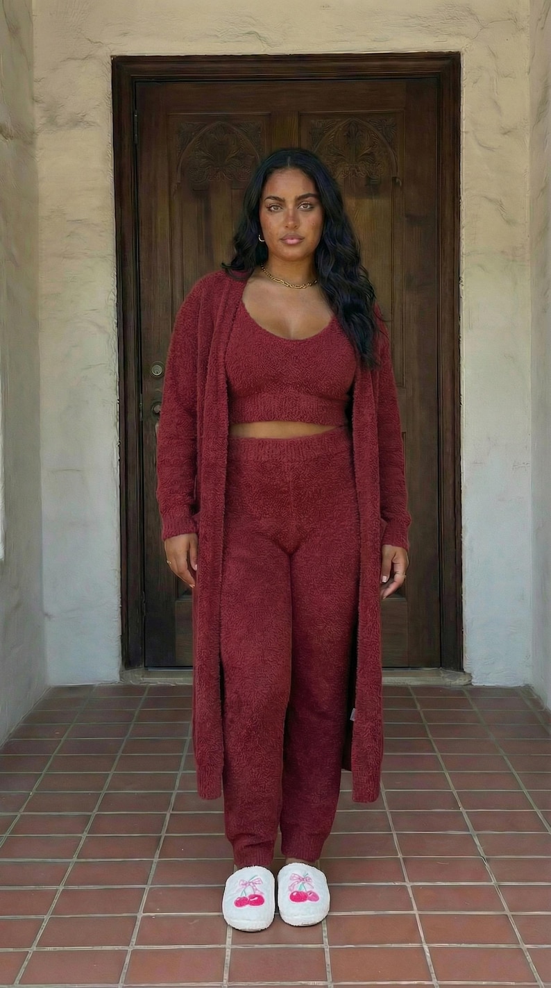 May include: A burgundy three-piece loungewear set, including a crop top, high-waisted trousers, and a long cardigan. The model is wearing white slippers with a cherry design. The outfit is made of a soft, textured material.