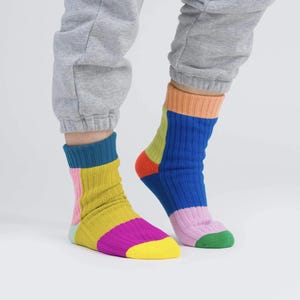 May include: A pair of colorful socks with a ribbed texture. The socks feature a patchwork design with sections of yellow, pink, blue, green, and orange. The socks are worn with gray sweatpants.