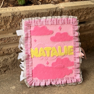 May include: A rectangular felt book cover in light pink featuring dark pink cloud appliques, small star shapes, and the name NATALIE in yellow felt letters. The border is finished with a pink and white gingham ruffled fabric trim.