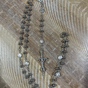 May include: A silver rosary with a silver crucifix and round beads. The rosary is laid out on a wooden surface.