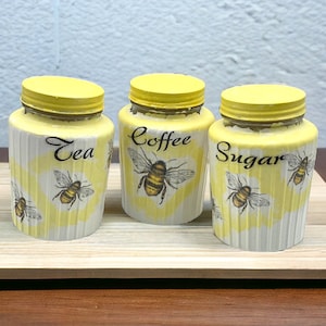 Può includere: Tre barattoli di ceramica gialli con disegni di api e scritte nere. I barattoli sono etichettati "Tè", "Caffè" e "Zucchero".