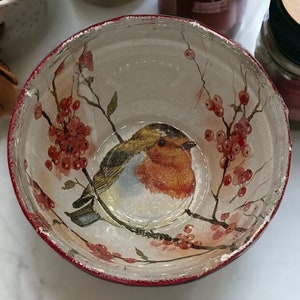 Może przedstawiać: Ozdobna ceramiczna miska z czerwonym obramowaniem i wzorem ptaków i jagód. Wnętrze przedstawia szczegółową ilustrację rudzika siedzącego na gałęzi z czerwonymi jagodami. Miska jest biała z postarzanym wykończeniem.