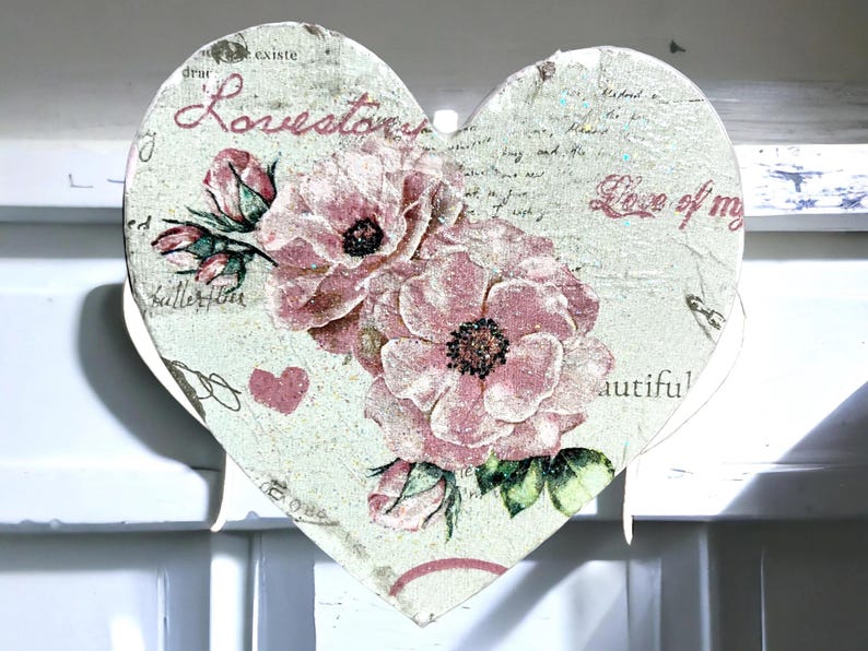 A heart-shaped decoration features a textured, glittery surface adorned with an intricate floral design. Soft pink blossoms intertwine with green leaves, creating a delicate, vintage aesthetic. Scattered text in varied fonts, including "Lovetown" and "Love of my," overlays the pattern, blending into the composition. The close-up reveals fine details, such as subtle shading and the embossed texture, enhancing the tactile and visual depth of the piece