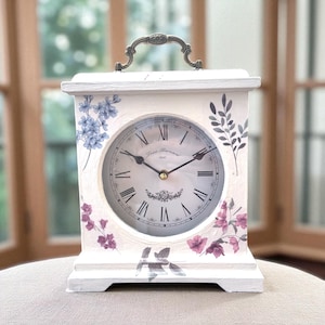 Puede incluir: Reloj de chimenea blanco con un diseño floral en morado y azul. La esfera del reloj tiene números romanos y un asa decorativa. El reloj es un elemento decorativo para el hogar.